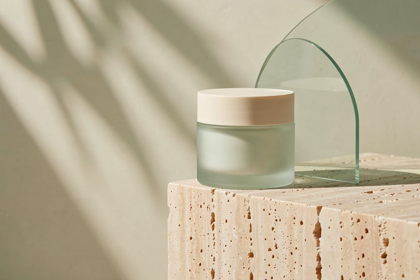 A frosted pale green skincare jar on sculptural ivory stone with soft botanical shadows.